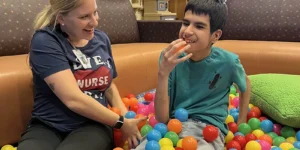 A nurse and child sit together in a colorful ball pit while the child holds an orange ball.