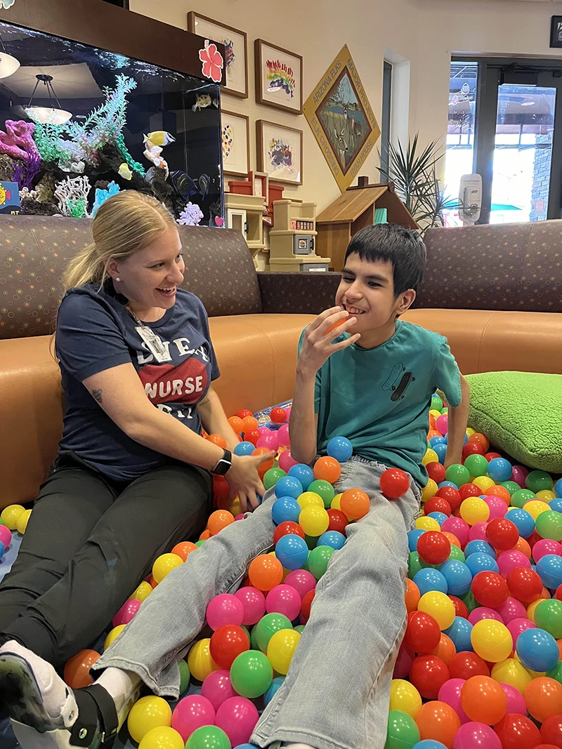 A nurse and child sit together in a colorful ball pit while the child holds an orange ball.